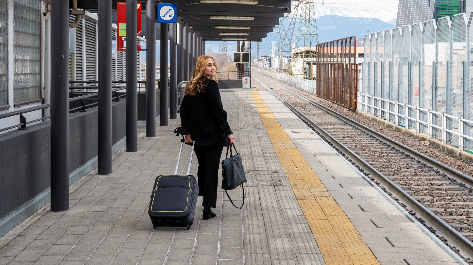 A woman with a bag and a suitcase is walking down the train platform. She is looking towards the rail tracks.