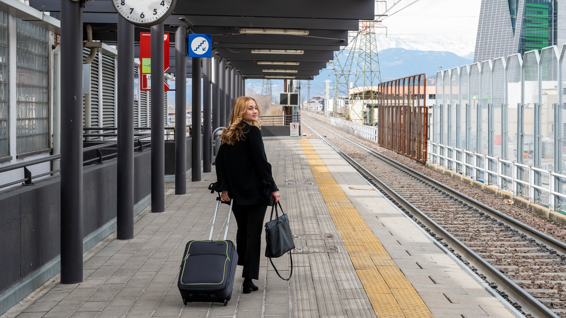 Una ragazza con borsa e valigia cammina lungo la banchina della stazione. Guarda verso i binari.